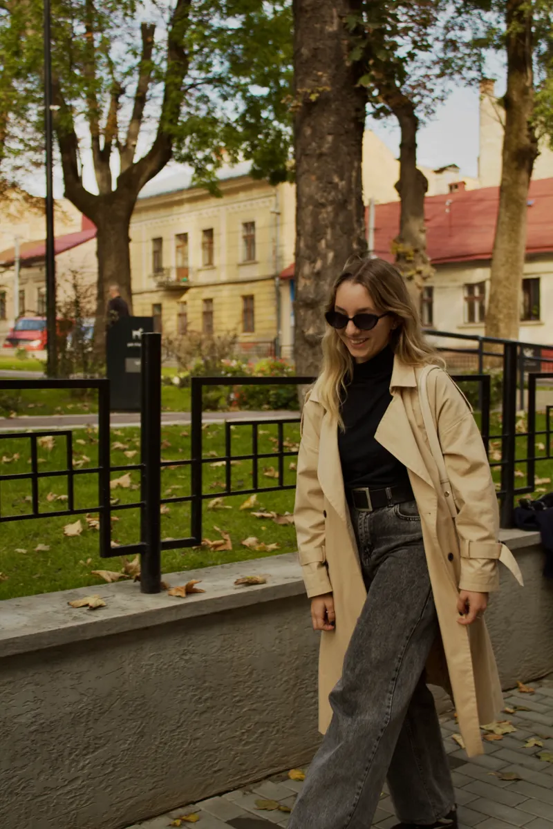 A smiling woman in sunglasses walking past railings in a beige trench coat, black polo neck and grey wide leg jeans
