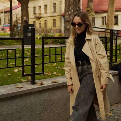 A smiling woman in sunglasses wearing a beige trench coat over a black polo neck and grey jeans