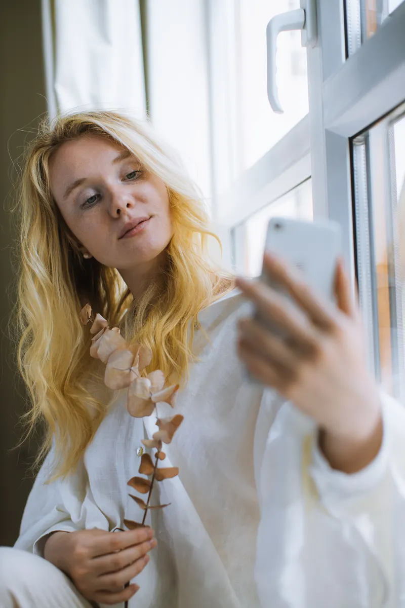 A blonde woman in a white shirt taking a selfie beside a bright window