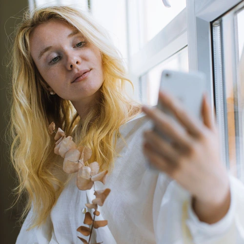 A blonde woman in a white shirt taking a selfie beside a bright window