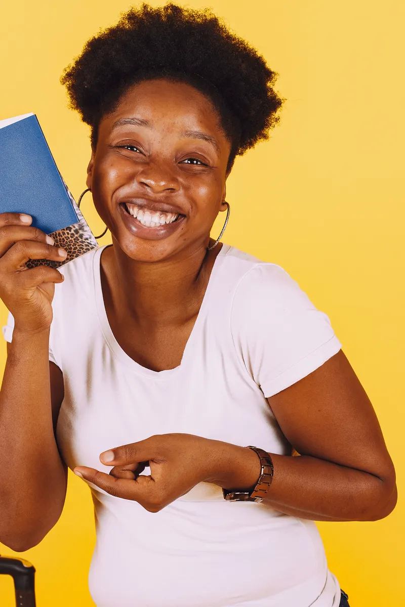 A laughing woman in a white T-shirt holding up a blue passport against a yellow wall