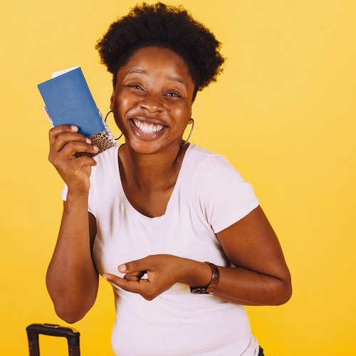 A laughing woman in a white T-shirt holding up a blue passport against a yellow wall